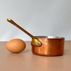 Pode incluir: Uma pequena panela de cobre com uma alça de latão e um ovo castanho. A panela tem um exterior de cobre brilhante e um interior prateado. A alça é fixada com rebites. O texto "MADE IN FRANCE" é visível.
