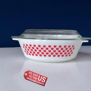 May include: White Pyrex casserole dish with a clear glass lid. The dish has a pattern of red tulips. The dish has a rectangular shape with rounded corners and side handles. A tag that says "WE SHIP TO THE US GIFTS = NO TARIFFS" is in the image.