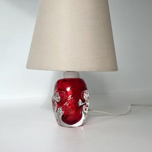 May include: A decorative table lamp with a red glass base and a beige lampshade. The base has a unique, textured design. The lamp is plugged in and ready to use. A stylish addition to any home decor.