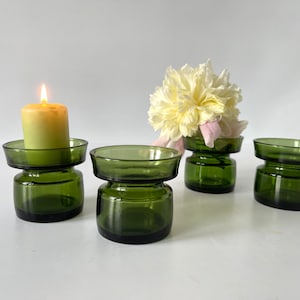 Può includere: Quattro portacandele in vetro verde, uno con una candela gialla accesa e un altro con fiori bianchi e rosa. I portacandele hanno un design a due livelli, con una base rotonda e un piatto poco profondo sopra.