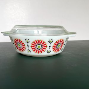 May include: A white glass casserole dish with a clear glass lid and two side handles. The exterior features a repeating pattern of stylized red, black, and lime green floral bursts arranged in a horizontal band around the circular bowl.