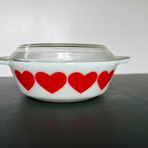 May include: A white glass casserole dish with a repeating pattern of bright red hearts around the exterior. The round bowl features two small side handles and comes with a clear, circular glass lid that rests securely on the top rim.