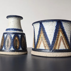 Può includere: Due vasi in ceramica con sfondo bianco e motivi geometrici blu e marroni. Il vaso a sinistra è più alto e più stretto del vaso a destra.
