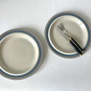 Op de afbeelding: Twee crèmekleurige keramische borden met blauwe en bruine rand. Eén bord heeft een vork en mes met zwarte handvatten en gouden accenten. Het servies staat op een witte ondergrond.