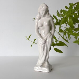Puede incluir: Una figura de cerámica esmaltada en blanco que representa a una persona de pie con el pelo largo, cubierta con una tela que envuelve la parte inferior del cuerpo. La escultura tiene una superficie lisa y brillante sobre una base rectangular.