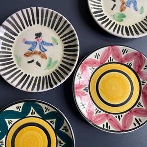 Op de afbeelding: Een verzameling handbeschilderde keramische borden. Eén bord toont een figuur in een blauwe jas en zwarte hoed, omringd door zwarte verticale lijnen. Andere borden hebben bloemen- en geometrische ontwerpen in geel, roze en groen.