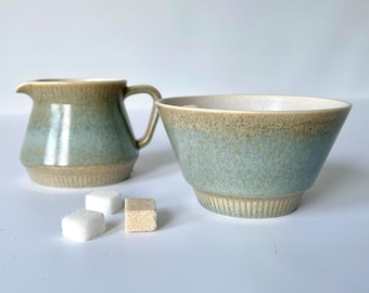 Może przedstawiać: Zestaw ceramiczny dzbanek do śmietanki i cukiernica w kolorze zielonym i brązowym. Dzbanek ma uchwyt i fakturowaną powierzchnię. Cukiernica jest okrągła i ma fakturowaną powierzchnię.