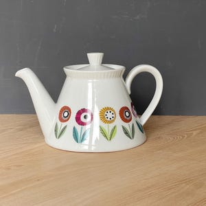 May include: White ceramic teapot with a lid and handle. The teapot is decorated with colorful floral designs in red, yellow, and purple. The teapot is sitting on a wooden surface.