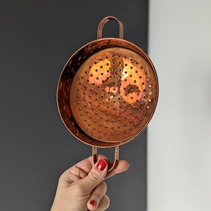 May include: A copper kitchen colander with a perforated bowl and two handles. The round colander is designed for draining liquids from food. The copper has a shiny, reflective surface. The colander's diameter is approximately 20 cm.