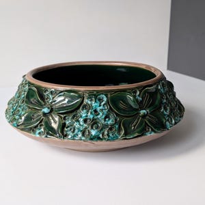 Puede incluir: Un cuenco de cerámica verde con un diseño floral en relieve. El cuenco tiene un borde ancho y un interior profundo.