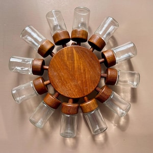 Puede incluir: Un estante de especias de madera con 12 tarros de vidrio transparentes. Los tarros están dispuestos en un círculo alrededor de una plataforma central de madera. El estante está hecho de madera oscura y tiene un aspecto rústico.