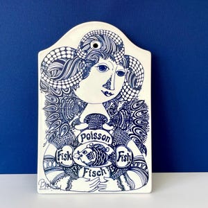 Puede incluir: Placa de pared de cerámica blanca con una ilustración azul de una mujer sosteniendo un pez. La placa tiene las palabras "Poisson", "Fisk" y "Fisch" alrededor del pez. El cabello y la ropa de la mujer están decorados con intrincados patrones azules.