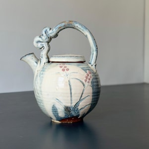 以下が含まれることがあります： 装飾的な結び目のディテールが施された高いアーチ型のハンドルを持つ、丸い陶器のティーポット。本体はオフホワイトで、青灰色の筆致と細い茎に描かれたピンクのベリーの房が特徴です。一致する蓋と短い注ぎ口が付いています。