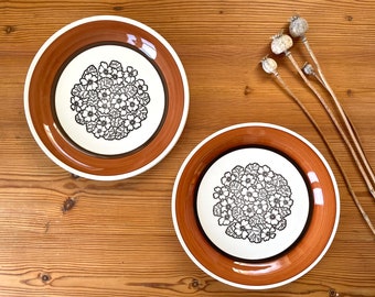 以下が含まれることがあります： 黒の花柄が描かれた茶色と白のセラミック製ディナー皿2枚。皿は茶色の縁取りと白の中央に黒の花柄が描かれています。