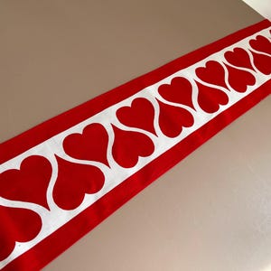 May include: Red and white table runner with a repeating pattern of white hearts on a red background.