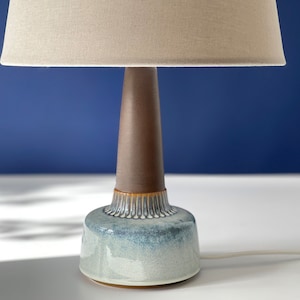 Puede incluir: Una lámpara de mesa de cerámica con una pantalla de tela beige. La base de la lámpara es una combinación de cerámica esmaltada azul y blanca, con una columna central marrón y cónica. La lámpara está sobre una superficie blanca sobre un fondo azul.