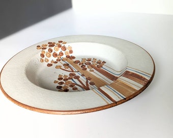 Op de afbeelding: Een witte keramische kom met een bruin en blauw ontwerp. De kom heeft een brede rand en een ondiepe put. Het ontwerp toont gestileerde bomen met bruine takken en bruine en blauwe bladeren.
