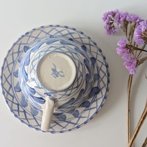 Puede incluir: Vista superior de un juego de taza y platillo de cerámica con un diseño azul y blanco. La taza está boca abajo sobre el platillo. Una ramita de flores moradas está a la derecha de la taza y el platillo.