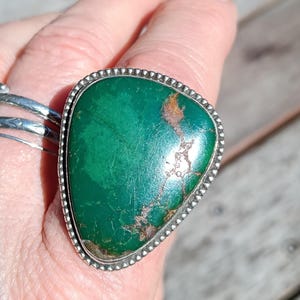 May include: A large, triangular, polished green stone ring set in a silver bezel with a beaded edge. The stone features natural brown matrix inclusions. A secondary, thinner silver band with etched detailing is worn on the same finger.