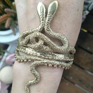 May include: A gold-colored bracelet featuring a design of two snakes with their heads facing upwards. The snakes' bodies wrap around the wrist, creating a unique and eye-catching accessory. The bracelet is made of a shiny metal.