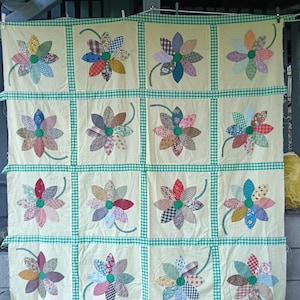 May include: A patchwork quilt featuring a grid of sixteen square blocks on a pale yellow background. Each block contains a flower appliqué with multi-coloured patterned fabric petals, a solid green centre, and a green stem, separated by green gingham sashing.