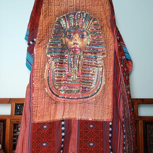 May include: A decorative textile featuring a detailed depiction of a pharaoh's mask, adorned with colorful sequins and beads. The textile has a warm, earthy color palette with intricate patterns and hieroglyphic-style text. The overall design evokes an Egyptian theme.