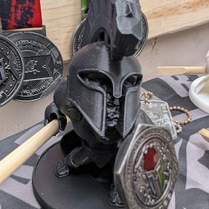 May include: A black 3D-printed figurine of a Spartan warrior wearing a crested helmet and armor. The figure holds a wooden spear in one hand and a metallic, hexagonal shield with red and green accents in the other, all mounted on a circular base.