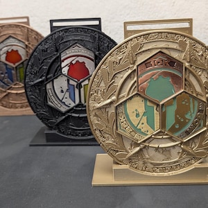 May include: Three circular medals displayed on stands, colored in gold, black, and bronze. Each medal features a central Spartan helmet emblem with text including 10K, 25K, and SPRINT 5K. The outer rims are decorated with intricate laurel leaf patterns.