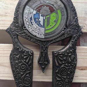 Könnte beinhalten: Dunkelgraue, spartanische Helmmedaille mit aufwendigen floralen und geometrischen Mustern. In der Mitte befindet sich ein kreisförmiges Emblem mit dem Text "SPARTAN 2024" und "TRIFECTA TRIBE" um eine segmentierte Grafik in Blau, Grün und Grau.