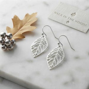 May include: A pair of silver-toned drop earrings featuring a cut-out leaf design with curved hooks. Beside the earrings is a small cluster of metallic silver spheres and a tan dried oak leaf, with a white card printed with WALK IN THE WOODS.