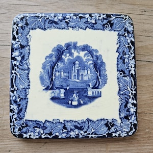 May include: Square ceramic coaster with a blue and white design. The centre features a detailed landscape scene with a building, trees, and figures. The border is adorned with a blue floral and leaf pattern. The coaster is set on a wooden surface.