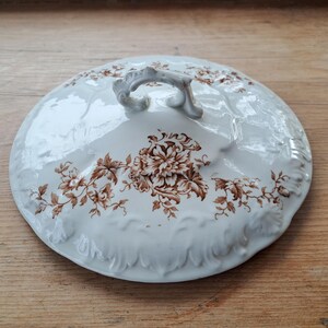 May include: A white ceramic lid with a brown floral pattern and a decorative handle. The lid has a glossy finish and a raised, textured edge. The floral design features large flowers and leaves in shades of brown. This lid is likely for a serving dish or casserole.