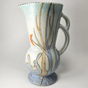 May include: A ceramic jug in light blue and white, decorated with a hand-painted design of trees and rabbits. The jug has a curved handle and a decorative rim. The design includes orange, brown, and blue accents. The jug is a decorative item.