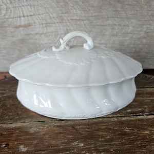 May include: A white ceramic lidded dish with a shell-like design. The lid has a curved handle and a scalloped edge. The dish has a textured, ribbed body and sits on a wooden surface.