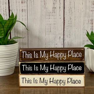 May include: Three wooden signs stacked on top of each other. Each sign is painted a different color: brown, black, and white. The signs all have the same text: "This Is My Happy Place" in white lettering.