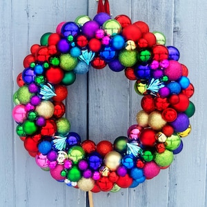 May include: A vibrant Christmas wreath composed of various sized ornaments in red, green, blue, purple, pink, and gold. The wreath is adorned with small, decorative blue accents and gold bells, creating a festive holiday decoration.