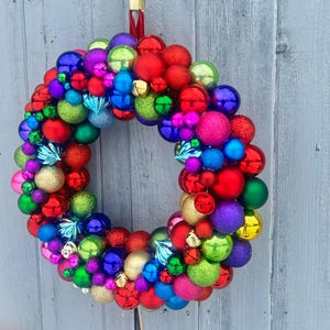 May include: A vibrant Christmas wreath composed of various sized ornaments in red, green, blue, purple, and gold. The wreath is hung against a weathered gray wooden background, creating a festive holiday decoration.