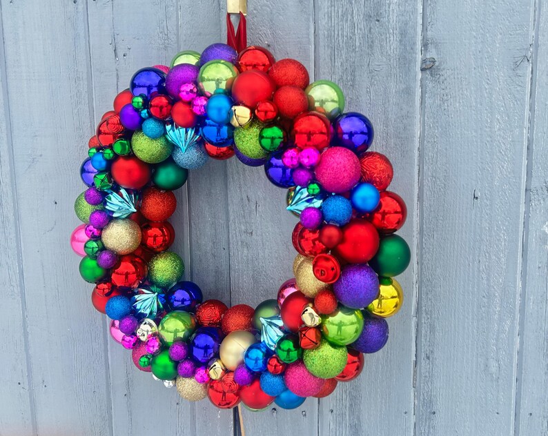May include: A vibrant Christmas wreath composed of various sized ornaments in red, green, blue, purple, and gold. The wreath is hung against a weathered gray wooden background, creating a festive holiday decoration.