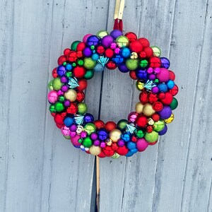 May include: A colorful Christmas wreath composed of various sized ornaments in red, green, blue, purple, and gold. The wreath is hanging against a gray wooden background, creating a festive holiday decoration.