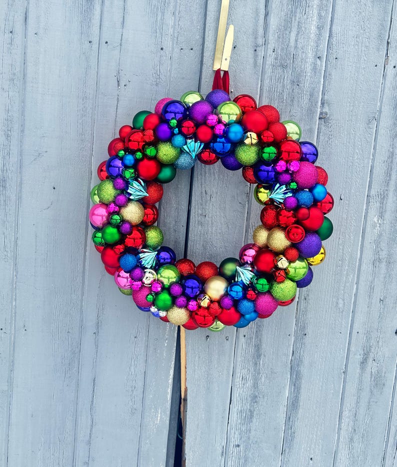 May include: A colorful Christmas wreath composed of various sized ornaments in red, green, blue, purple, and gold. The wreath is hanging against a gray wooden background, creating a festive holiday decoration.