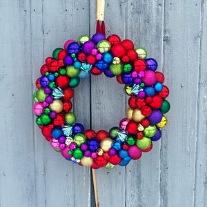May include: A vibrant Christmas wreath composed of various colorful ornaments in shades of red, green, blue, purple, pink, and gold. The wreath is suspended against a weathered gray wooden background, creating a festive holiday decoration.