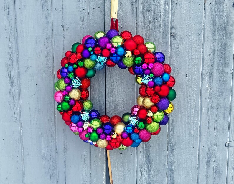 May include: A vibrant Christmas wreath composed of various colorful ornaments in shades of red, green, blue, purple, pink, and gold. The wreath is suspended against a weathered gray wooden background, creating a festive holiday decoration.