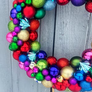 May include: A colorful Christmas wreath composed of various sized ornaments in red, green, blue, purple, gold, and pink. The wreath is arranged in a circular shape, with a variety of textures and finishes, including shiny, matte, and glitter.