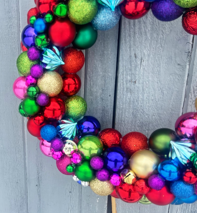 May include: A colorful Christmas wreath composed of various sized ornaments in red, green, blue, purple, gold, and pink. The wreath is arranged in a circular shape, with a variety of textures and finishes, including shiny, matte, and glitter.