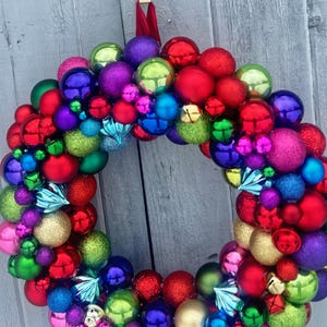 May include: A colorful Christmas wreath made of various sized ornaments in red, green, blue, purple, and gold. The wreath is round and has a red ribbon for hanging. The ornaments have a shiny, reflective surface.