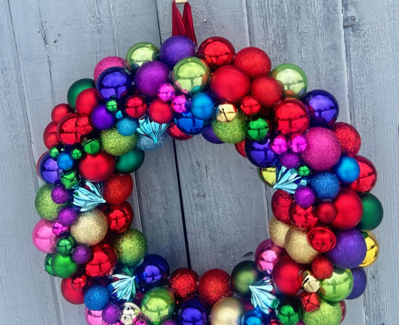 May include: A colorful Christmas wreath made of various sized ornaments in red, green, blue, purple, and gold. The wreath is round and has a red ribbon for hanging. The ornaments have a shiny, reflective surface.