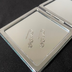 Puede incluir: Un par de pendientes plateados con el símbolo del infinito y ganchos curvos. Los pendientes se exhiben sobre una superficie reflectante dentro de un espejo compacto con marco plateado, sobre un fondo negro.