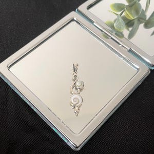 Puede incluir: Un colgante de plata con dos incrustaciones circulares de concha de ojo de Shiva engastadas en un marco de metal pulido. El diseño incluye una pequeña anilla en la parte superior para una cadena, con las conchas mostrando un patrón en espiral.