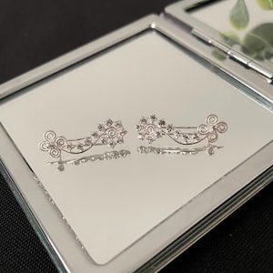 Op de afbeelding: Een paar zilverkleurige oorcrawlers met een bloemmotief. Elke oorbel heeft een gebogen staaf met kleine, heldere kristallen en decoratieve krullen. De oorbellen worden gepresenteerd op een spiegeloppervlak.