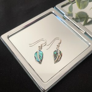 May include: A pair of silver leaf earrings with iridescent abalone shell inserts. The earrings are displayed on a silver compact mirror against a black background. The earrings have a hook closure.